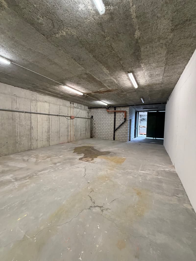 Empty concrete corridor with exposed pipes, a drain, and a small puddle of water on the floor.
