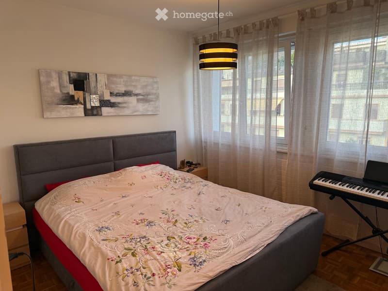 A well-lit bedroom with a large bed, a keyboard and laptop on a stand, a nightstand with small items, and a modern painting on the wall.
