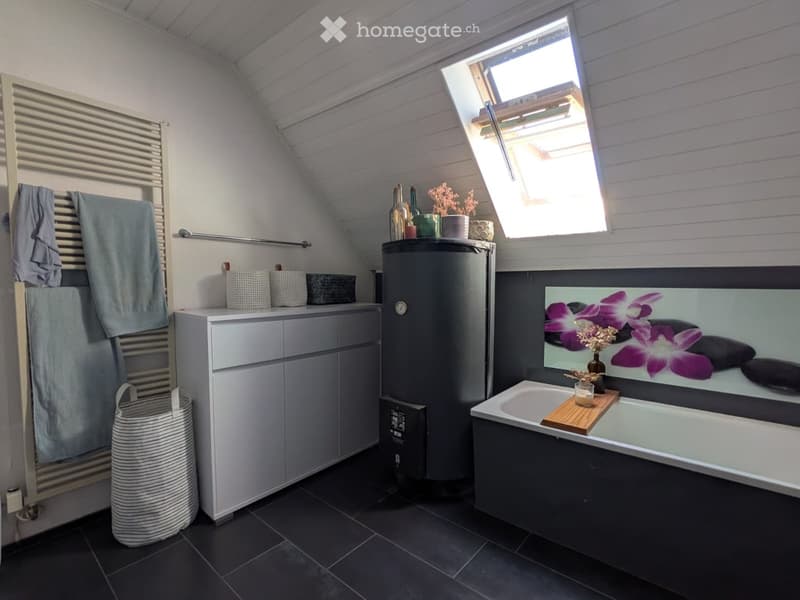 Modern bathroom with dark tiles, a bathtub, a cabinet, a towel rack, and decorative items including a mirror with a floral print and a small candle on the edge of the tub.