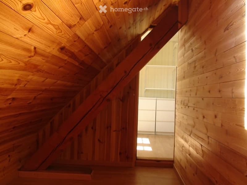 A narrow wooden hallway with exposed wooden beams and a slanted ceiling, leading to a bright, open space with white walls and a glass door.