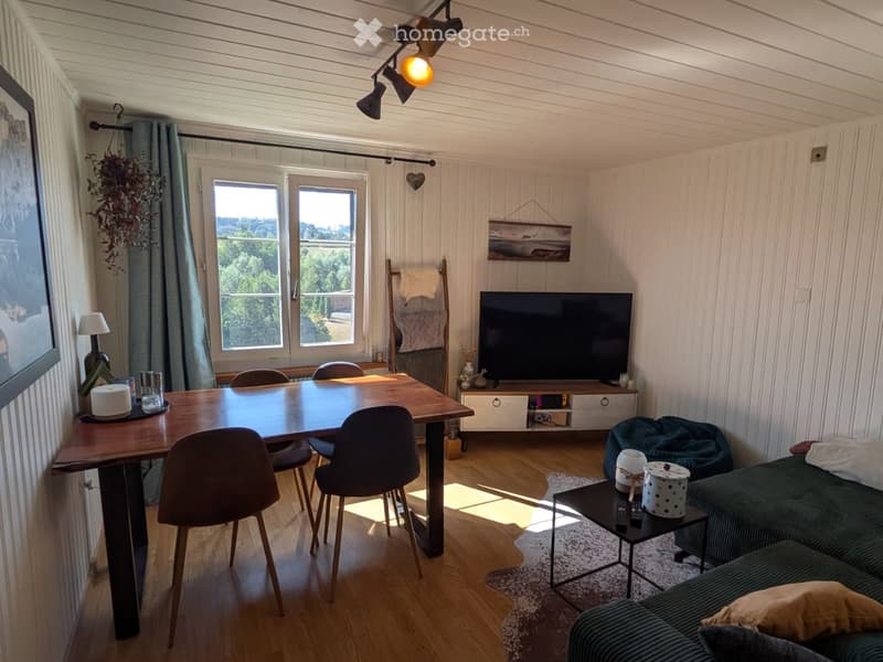 Bright living room with wooden flooring, large windows, a green sofa, coffee table, dining table with chairs, and a TV stand.