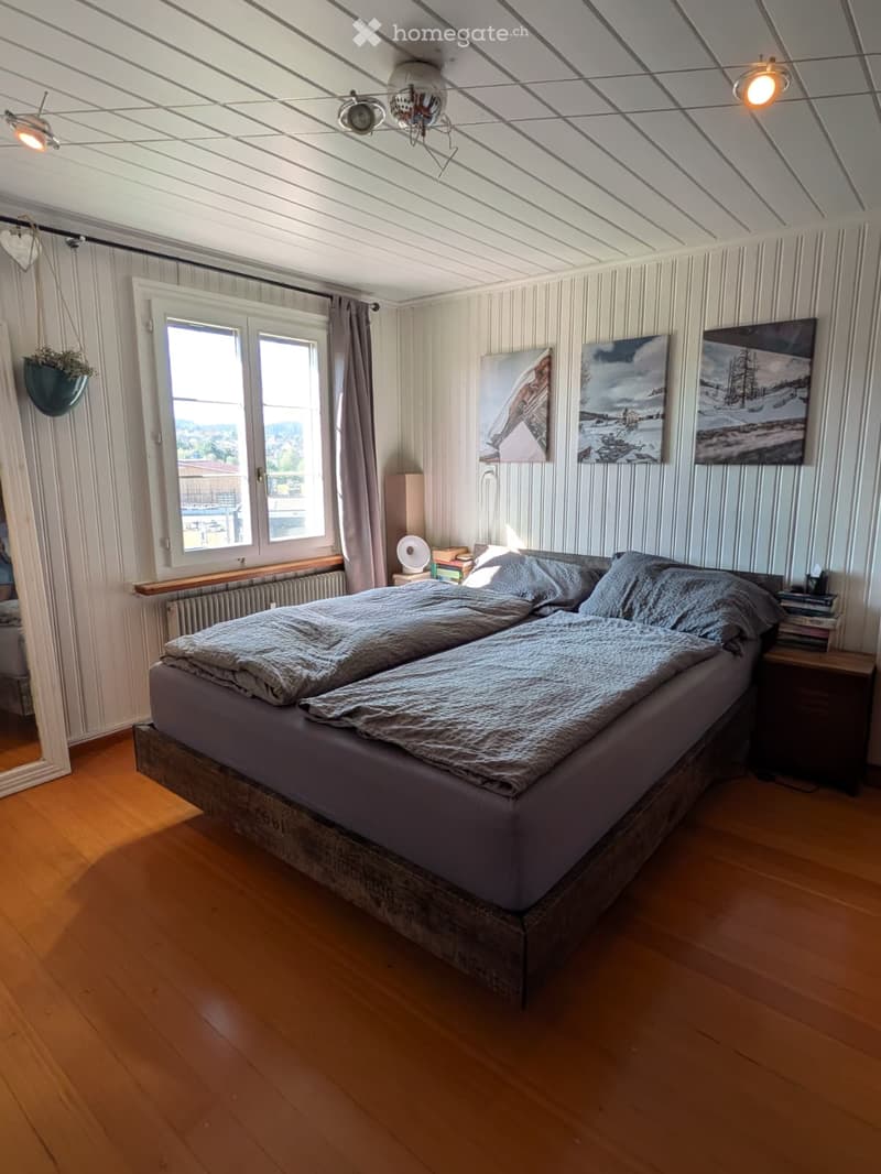Cozy bedroom with wooden flooring, a large bed with grey bedding, a nightstand with a lamp, a mirror, and several paintings on the walls. The room features large windows with curtains and a hanging plant.