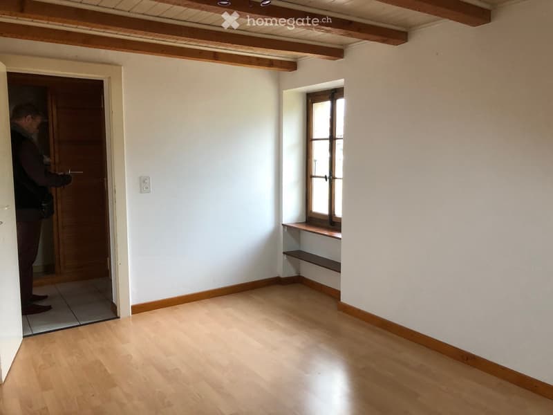 Empty room with wooden flooring, white walls, and a window with wooden frame. There is a small wooden bench under the window and a door leading to another room.