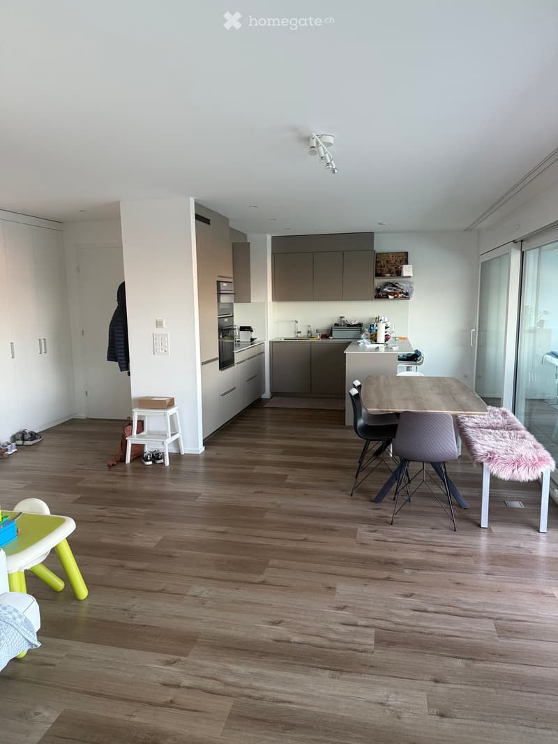 Open-plan living and dining area with modern kitchen, wooden flooring, and large windows. Includes a dining table with chairs, a small stool, a furry rug, and a child's table and chair.