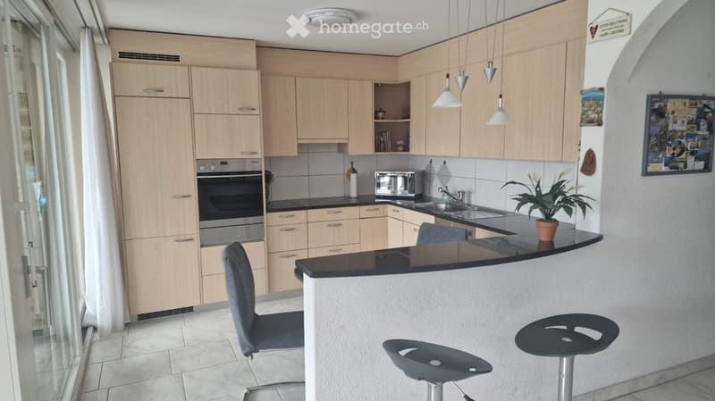 Modern kitchen with light wood cabinets, stainless steel appliances, black countertop, and bar stools.