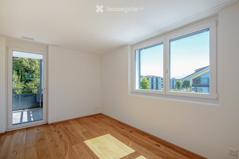 Empty room with wooden flooring, white walls, large windows, and a sliding glass door leading to a balcony.