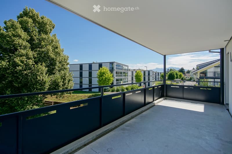 Modern balcony with a concrete floor, dark grey metal railing, and a covered roof. Offers a view of a contemporary residential complex and surrounding greenery.