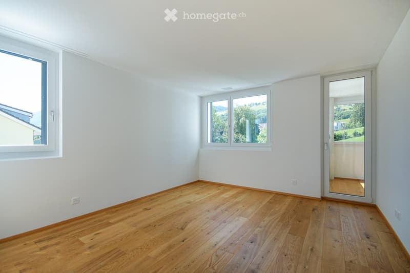 Empty room with light wooden flooring, white walls, and large windows offering a scenic view. The room features a sliding glass door leading to a balcony.