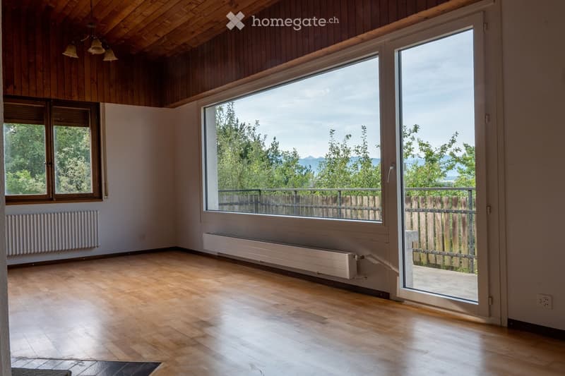 Bright, empty room with wooden flooring, large windows, and a sliding glass door leading to a balcony. The room features wooden paneling on the ceiling and walls, with radiators visible along the walls.