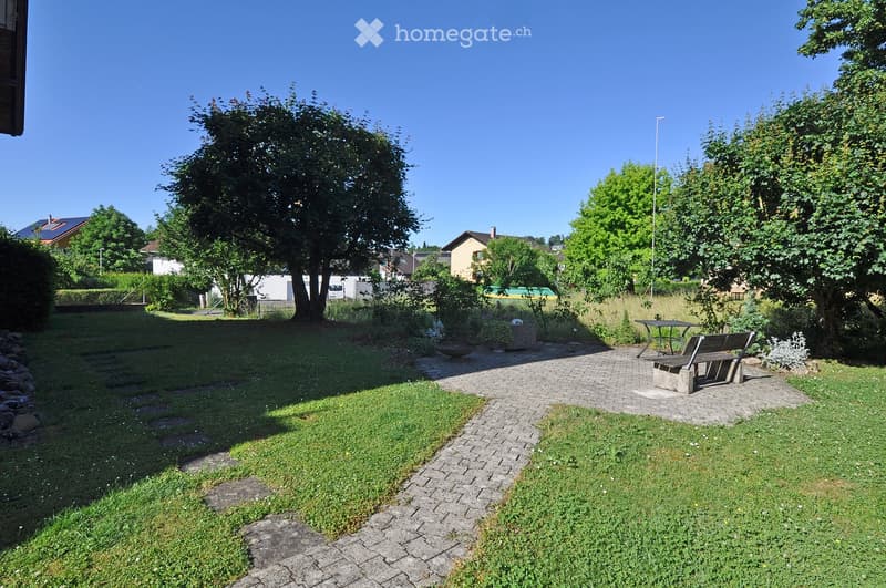 Well-maintained garden with a paved pathway, grassy areas, trees, a bench, and a small table, surrounded by neighboring houses.