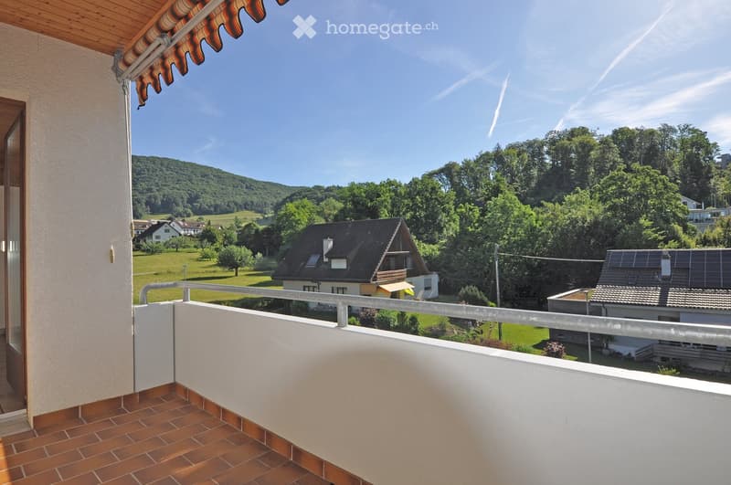 Balcony with wooden floor and white railing, overlooking a green landscape with trees and houses.