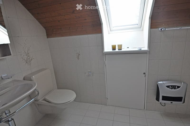 Bathroom with a toilet, sink, hand dryer, and a small window with two cups on the sill. The walls are tiled, and the ceiling is wooden.