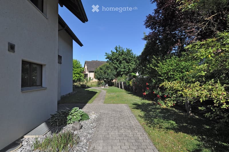 A well-maintained garden path leads to a house, surrounded by greenery and flowers, with a clear blue sky overhead.