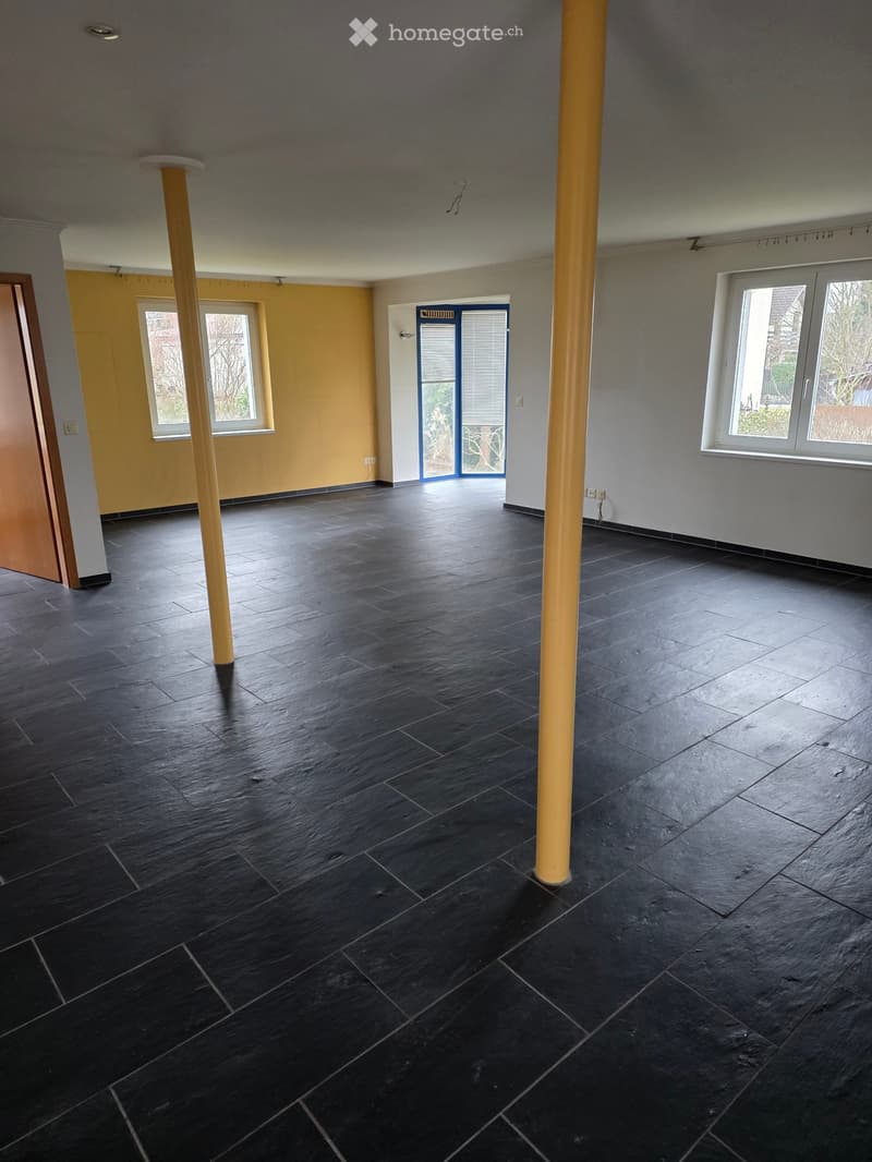 Empty room with dark tiled floor, two large windows, and a door leading to another area. The walls are painted in neutral tones with a yellow accent wall visible on one side.