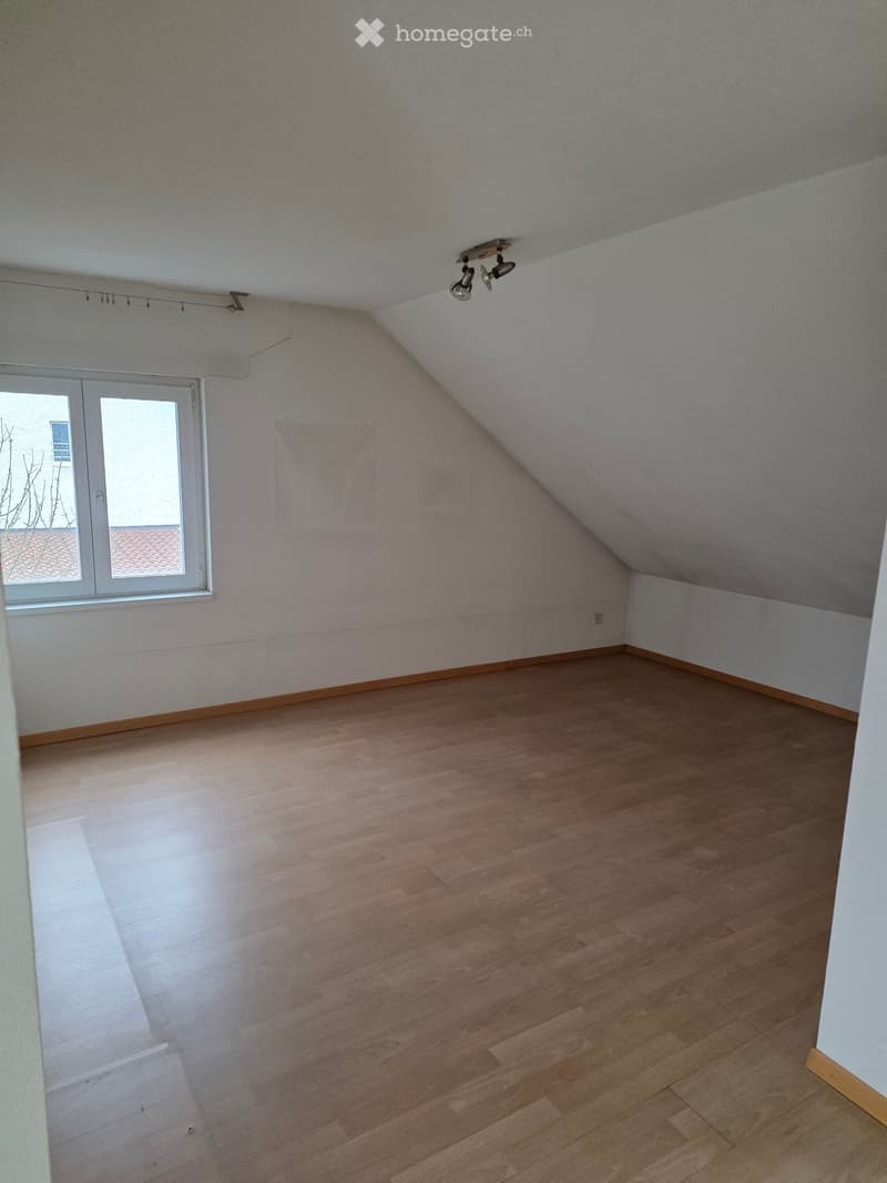 Empty attic room with wooden flooring, slanted ceiling, and a single window with white frame. No furniture or equipment present.