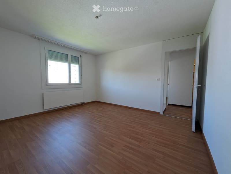 Empty room with wooden flooring, white walls, and a window with blinds. A radiator is visible below the window, and an open doorway leads to another space.