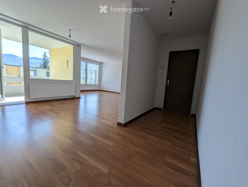Empty room with wooden flooring, white walls, large windows, and a door leading to another room.