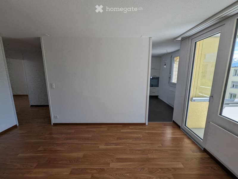 Empty room with wooden flooring, white walls, and large windows with blinds. The room is connected to a kitchen area visible in the background.