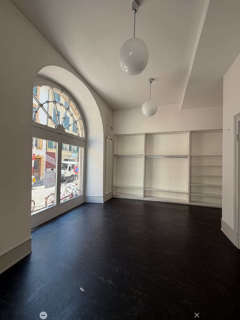 Empty room with dark flooring, white walls, built-in shelving unit, and large arched window with glass panes. Two modern pendant lights hang from the ceiling.