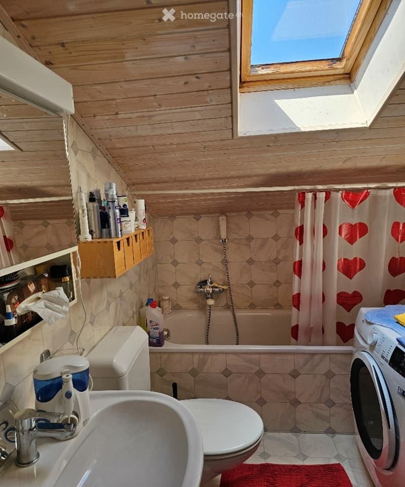 Bathroom with wooden ceiling, hexagonal tiles, sink, toilet, bathtub, shower curtain with heart pattern, washing machine, and various toiletries.
