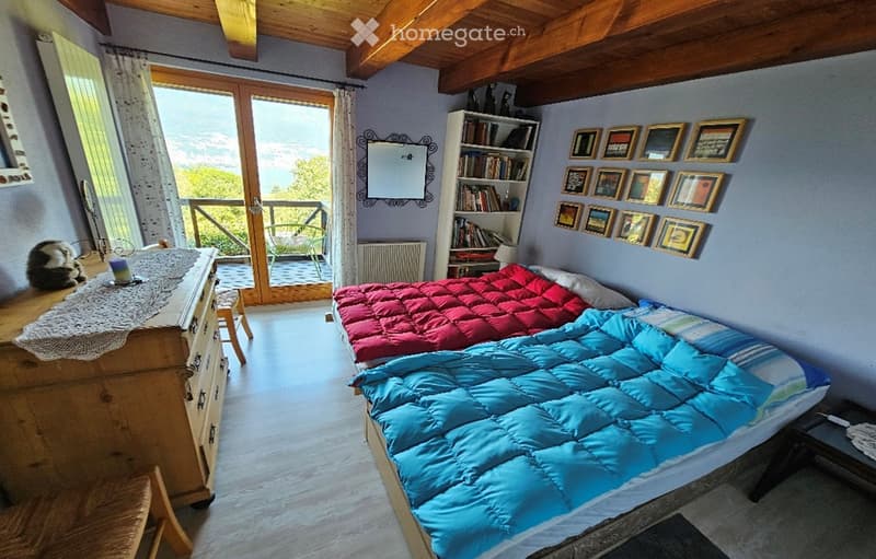 Cozy bedroom with wooden beams, large window with balcony access, double bed with blue and red duvet, nightstand, bookcase, mirror, and small table.