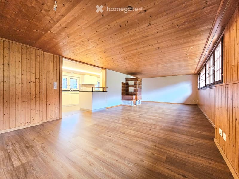 Empty room with wooden walls, ceiling, and floor, featuring large windows and an open doorway to another room with cabinets and a counter.