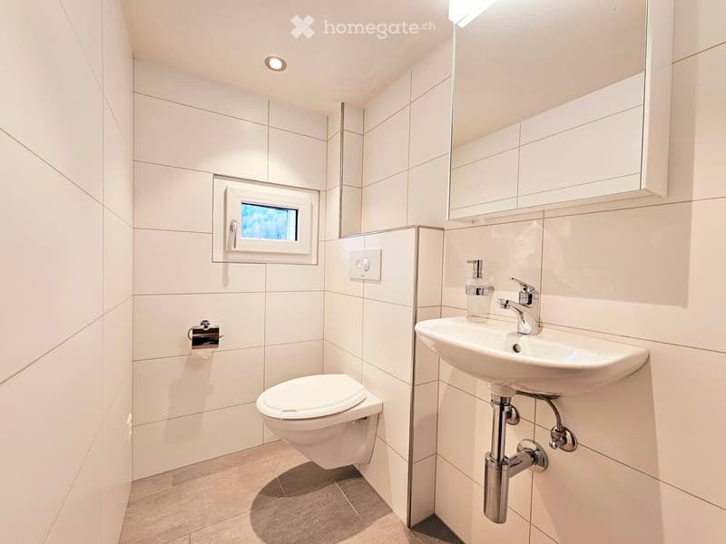 Modern bathroom with white tiles, a toilet, a sink with a chrome faucet, a mirror, a soap dispenser, and a small window.