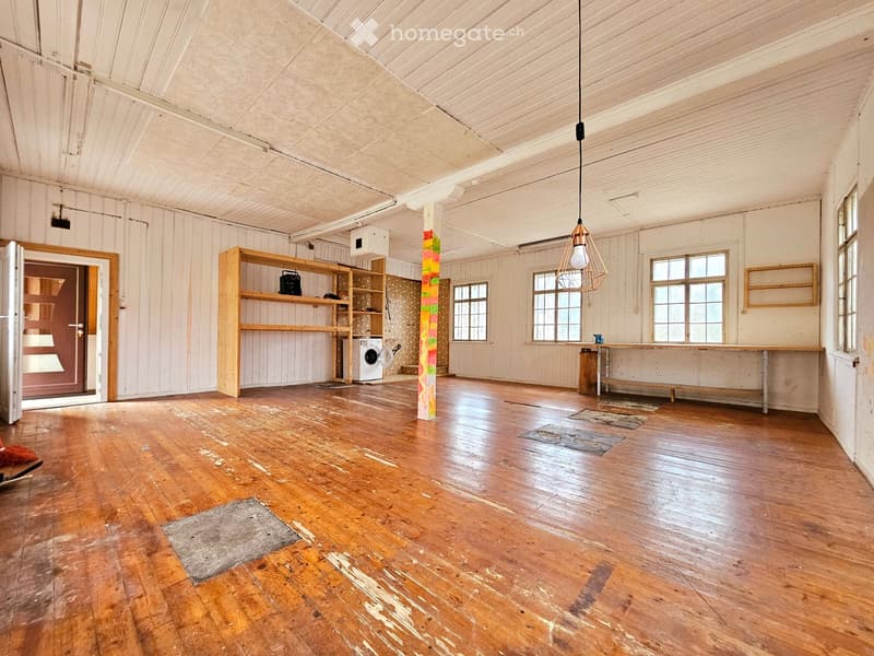 Large, open room with wooden floors, exposed wooden beams, and white walls. Features include a wooden shelf, a washing machine, a long wooden table, and a stool.