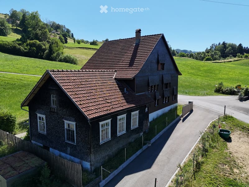 A two-story wooden house with a red-tiled roof, white-framed windows, and a small chimney, situated in a rural, green landscape with hills and trees in the background.