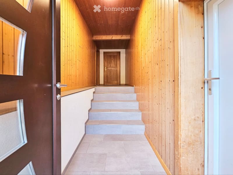 A narrow hallway with wooden panel walls, a staircase leading upwards, and a door at the end.
