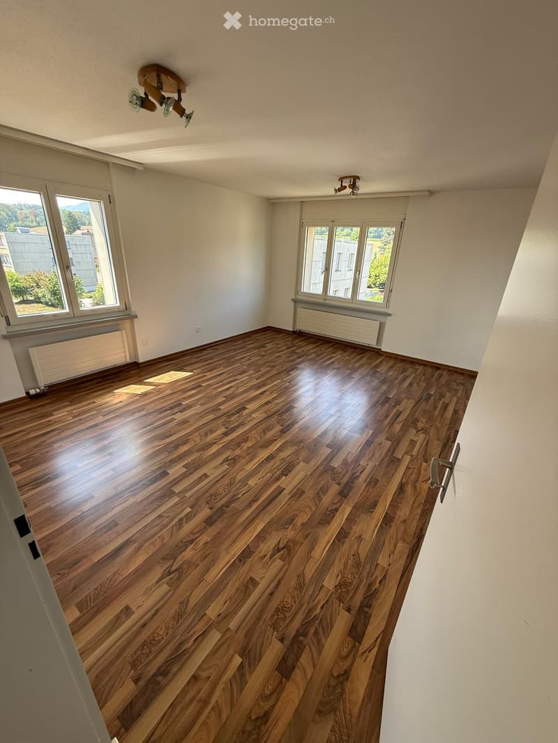 Empty room with wooden flooring, white walls, large windows, and ceiling lights.