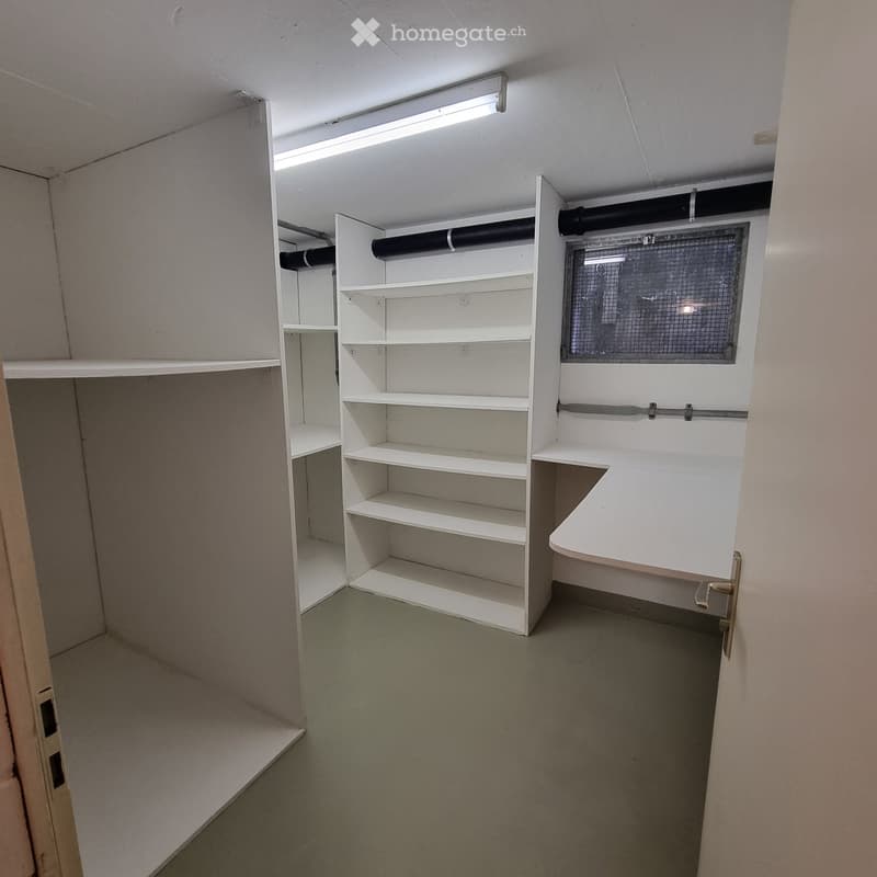 Empty room with white built-in shelving units, a small desk, and a window with a metal grid. The room has a concrete floor and white walls.