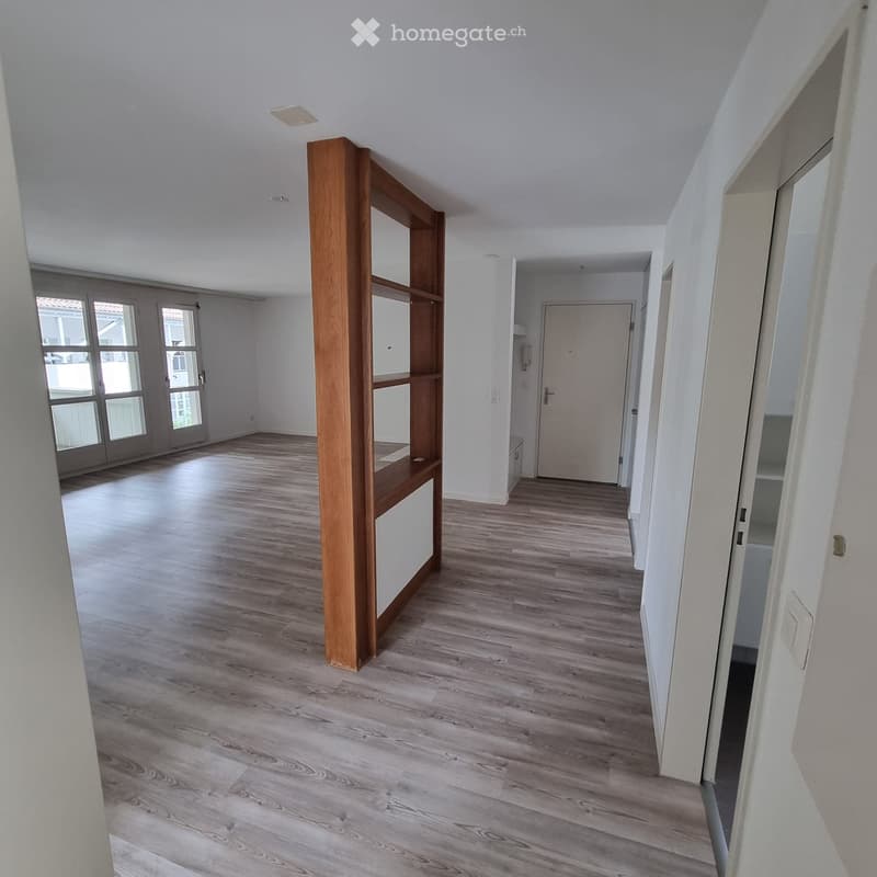 Empty hallway with light wooden flooring, white walls, and a wooden shelving unit on the left side. Natural light enters through a large window at the far end.