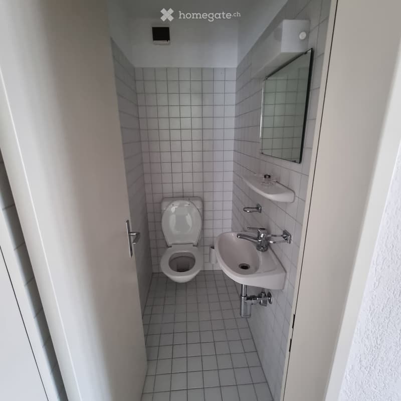 Small bathroom with white tiled walls and floor, featuring a toilet, sink, and mirror.