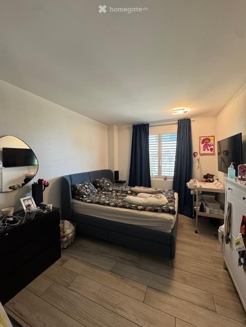 Cozy bedroom with wooden flooring, a large bed with floral bedding, a nightstand with a lamp, a mirror, and a small table with a chair near the window.