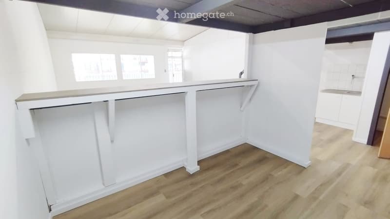 Empty room with white walls, wooden flooring, and a large white counter with built-in storage. Windows provide natural light.
