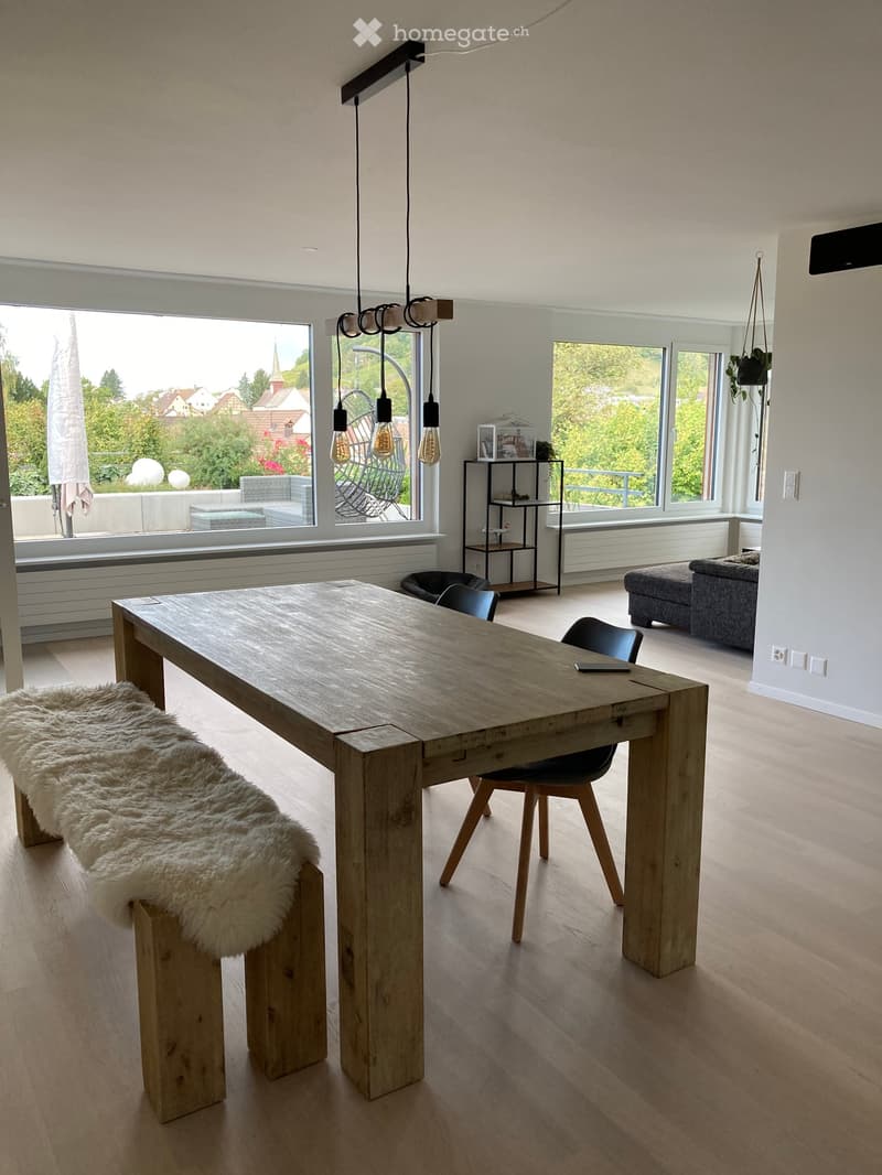 Bright, modern dining area with a large wooden table, bench, chairs, and a hanging lamp. Large windows offer a scenic view of the outdoors.