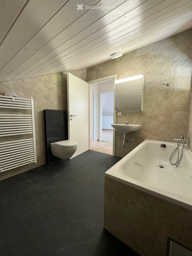Modern bathroom with dark tiled floor, white bathtub, sink, toilet, and radiator. Features a large mirror and an open door leading to another room.