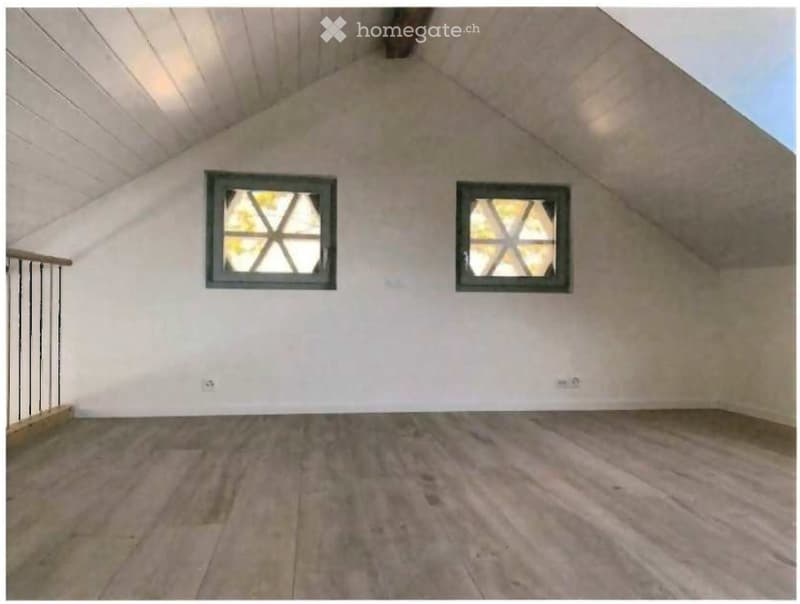 Empty attic space with wooden flooring, white walls, and two windows with decorative glass panes. The ceiling has exposed wooden beams and a sloped design.