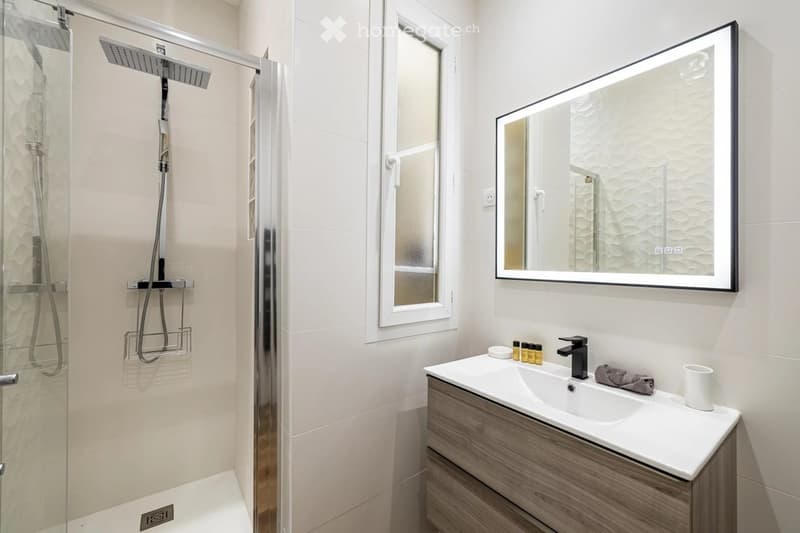 Modern bathroom with a glass-enclosed shower, a large illuminated mirror, a vanity with a sink, and a cup on the counter.