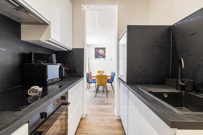 Modern kitchen with black countertops, stainless steel appliances, and a dining area with colorful chairs.