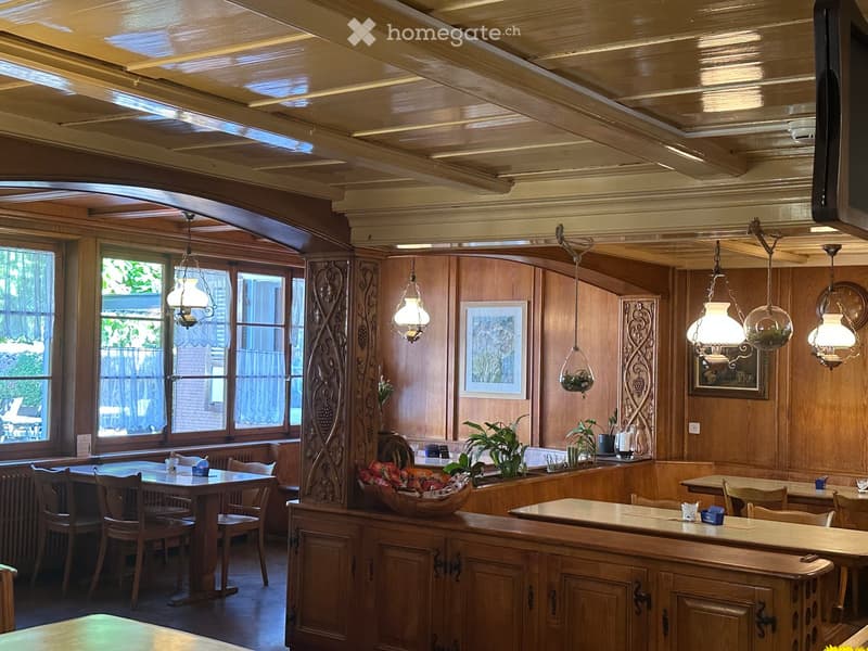 Cozy restaurant interior with wooden walls, exposed wooden ceiling beams, pendant lights, tables, chairs, and decorative elements.