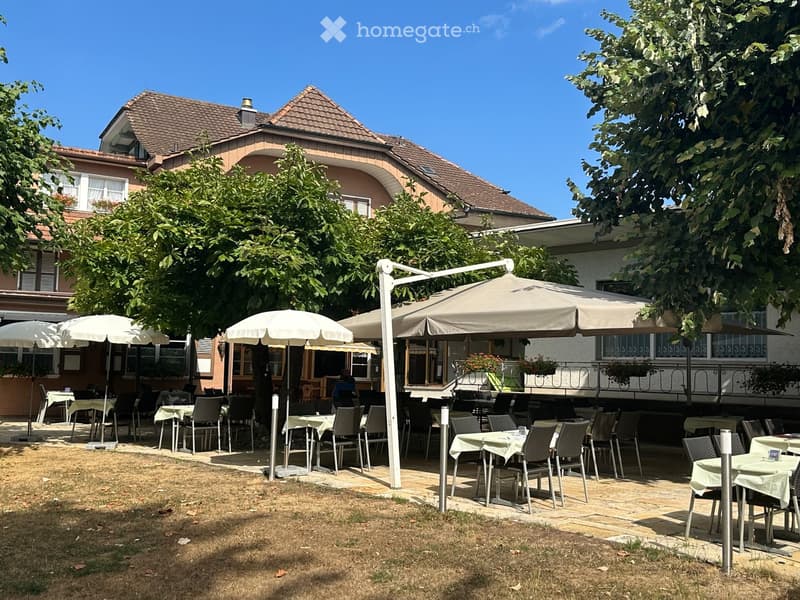 Outdoor seating area with tables, chairs, and umbrellas, surrounded by trees and a building with a tiled roof.