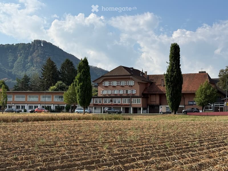 Two-story building with red-tiled roof and multiple windows, surrounded by trees and parked cars, with a grassy field in the foreground and mountains in the background.