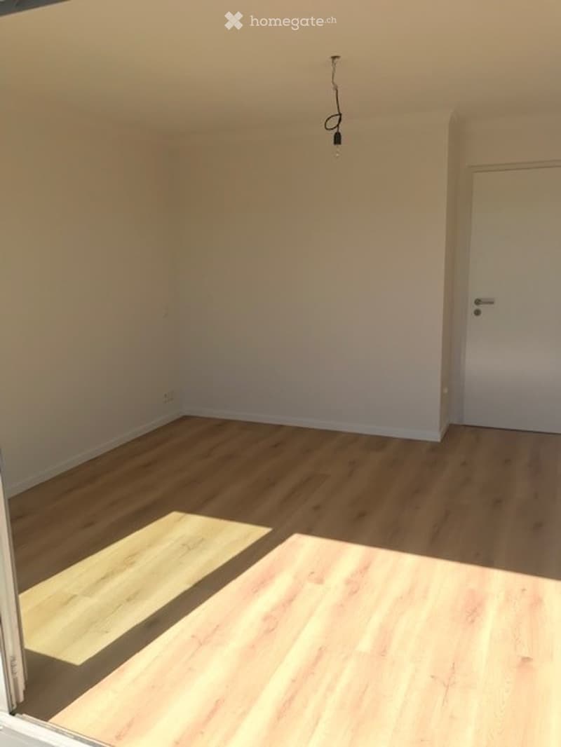 Empty room with light wooden flooring and white walls. A single door is visible on the right side, and a small section of a different flooring type is visible in the foreground.