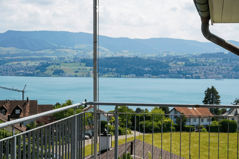 Balcony overlooking a scenic lake and distant mountains, with a metal railing and surrounding greenery.