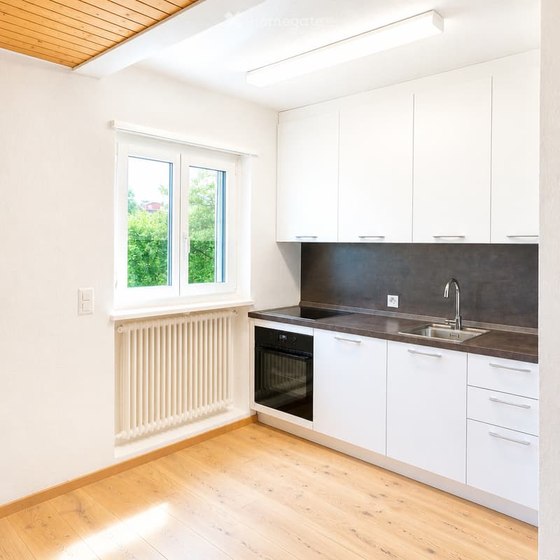 Modern kitchen with white cabinets, dark countertop, stainless steel sink and faucet, built-in oven, wooden flooring, and large windows.