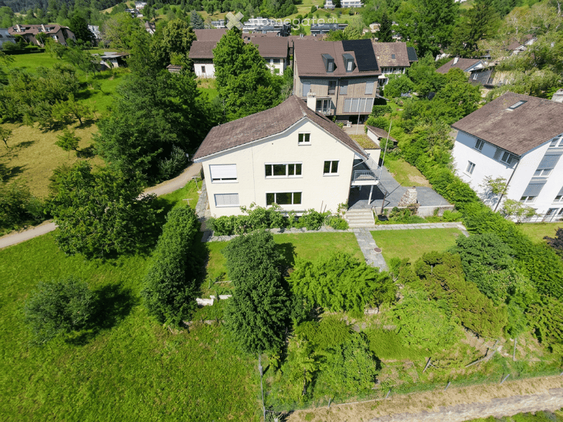 Aerial view of a two-story house with a well-maintained garden, surrounded by trees and neighboring houses in a hilly landscape.