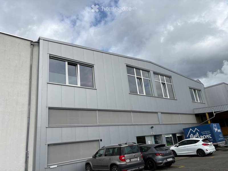 Modern industrial-style building with large windows and a flat roof, featuring several parked cars in front.