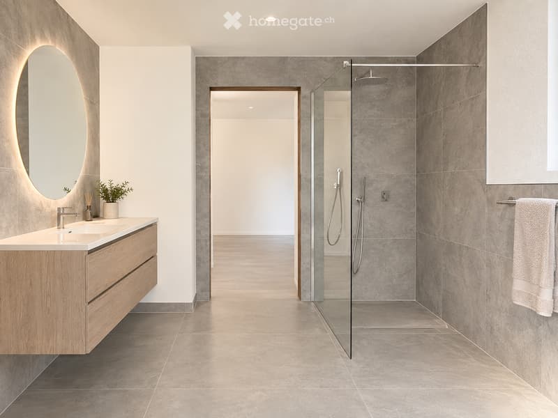 Modern bathroom with a wooden vanity, oval mirror with LED lighting, glass-enclosed shower, and a small potted plant on the counter.